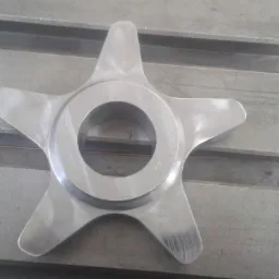 Metalowy element w kształcie gwiazdy z okrągłym otworem w środku, leżący na szarej, żłobionej powierzchni roboczej. Widoczne ślady obróbki na powierzchni metalu.