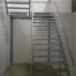 Metalowe schody drabiniaste z poręczą, prowadzące na antresolę w pomieszczeniu o panelowych ścianach.