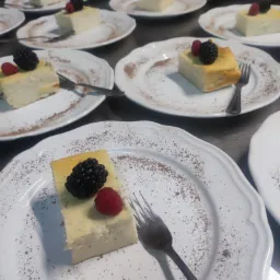 Porcje sernika nowojorskiego udekorowane maliną i jeżyną na białych talerzach z posypką kakaową, widoczne sztućce i przygotowanie do serwowania.