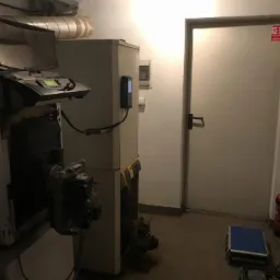 Wnętrze kotłowni z widocznym kotłem gazowym, rurami, skrzynką narzędziową i drzwiami przeciwpożarowymi w tle.