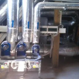 Trzy izolowane rury wentylacyjne z niebieskimi strzałkami na metalowej konstrukcji w pomieszczeniu technicznym z drewnianymi elementami i skrzynką elektryczną.