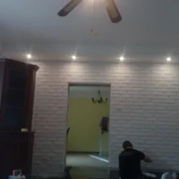 Mężczyzna w czarnej koszulce siedzi na podłodze, prawdopodobnie poprawiając wykończenie ściany z białej cegły w pokoju z oświetleniem punktowym i wiatrakiem sufitowym.