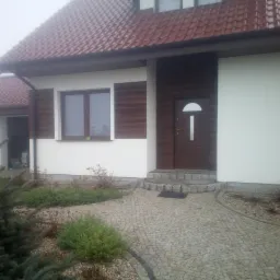 Gotowy dom jednorodzinny z brązowymi elementami elewacji, czerwoną dach&oacute;wką i kamiennym chodnikiem prowadzącym do wejścia.