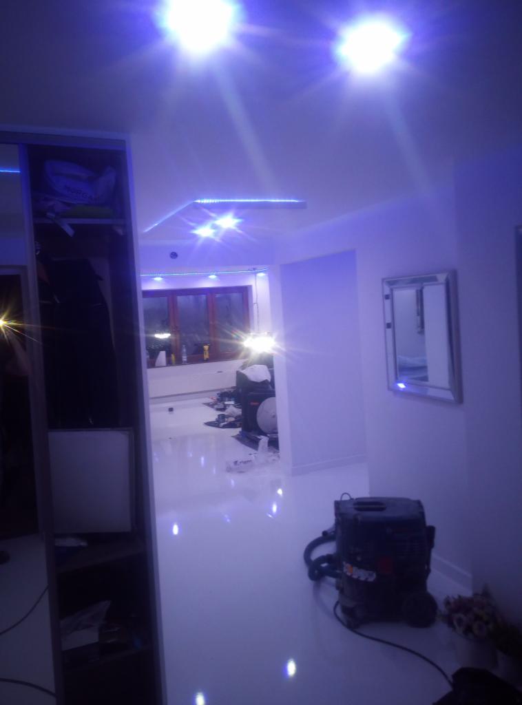 Wnętrze w trakcie prac wykończeniowych: błyszcząca, biała podłoga, widoczny odkurzacz przemysłowy, niebieskie oświetlenie LED na suficie, częściowo widoczna szafa wnękowa z półkami.