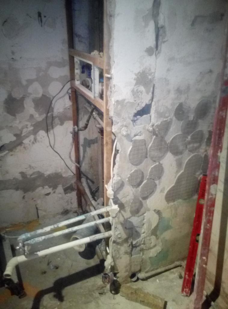 Niedokończony remont łazienki: widoczne rury, fragment ściany z pozostałościami po płytkach i siatce, drewniana konstrukcja pod zabudowę, poziomica oparta o ścianę.