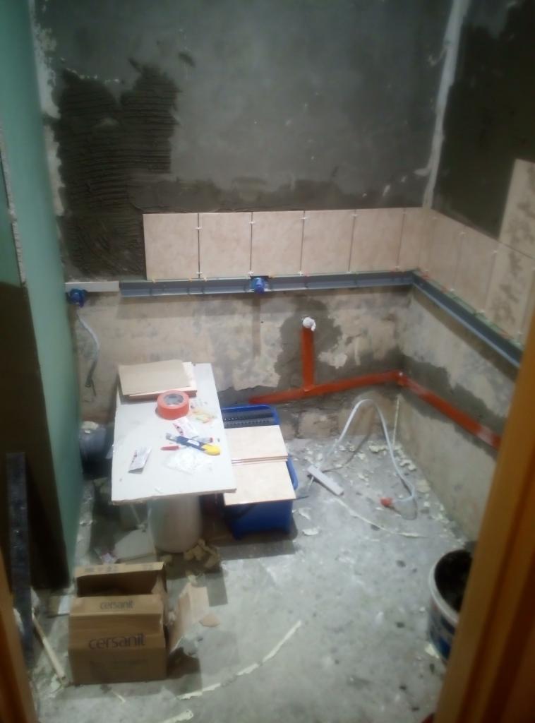 Wnętrze łazienki w trakcie układania płytek, widoczne narzędzia, rury instalacyjne i kartony po płytkach Cersanit.