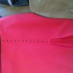 Czerwona sukienka z dekoracyjnymi czarnymi koralikami i plisowaniem, leżąca obok miarki krawieckiej z kwiatowym wzorem i skórzanej torebki na drewnianym stole.