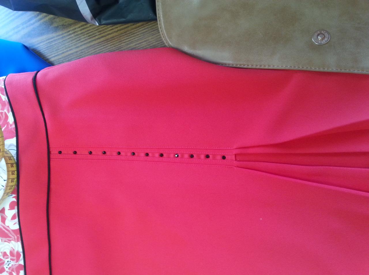 Czerwona sukienka z dekoracyjnymi czarnymi koralikami i plisowaniem, leżąca obok miarki krawieckiej z kwiatowym wzorem i skórzanej torebki na drewnianym stole.