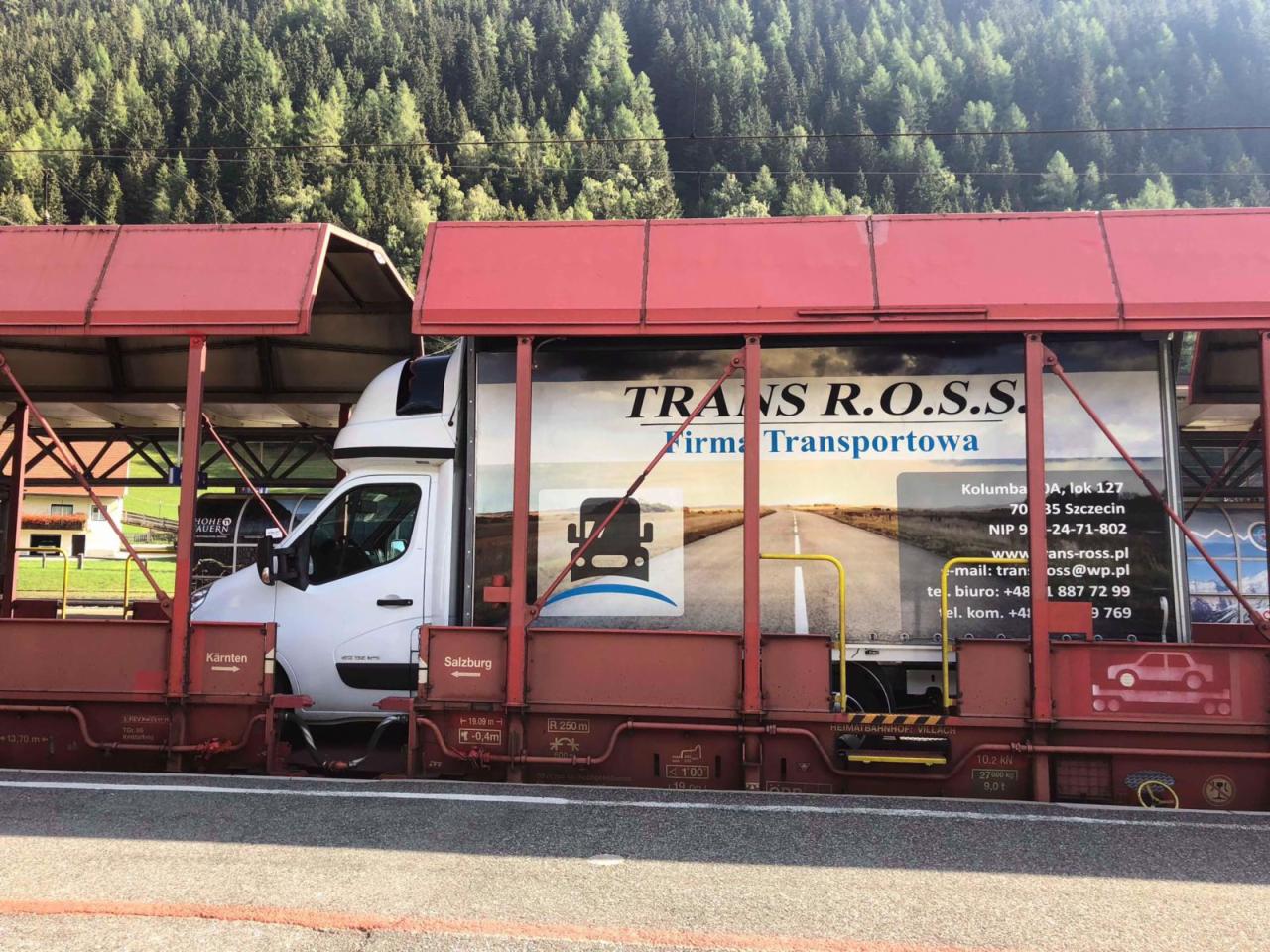 Biały bus transportowy firmy Trans R.O.S.S. załadowany na wagonie kolejowym, widok z boku, w tle zielone drzewa.