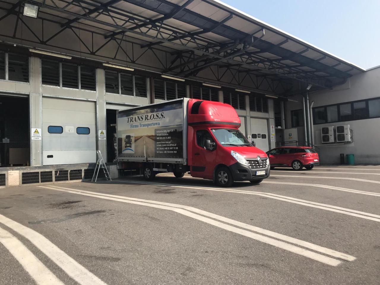 Czerwony bus transportowy z nadrukowanym logo firmy TRANS R.O.S.S. zaparkowany przed magazynem z rampą załadunkową i schodami.