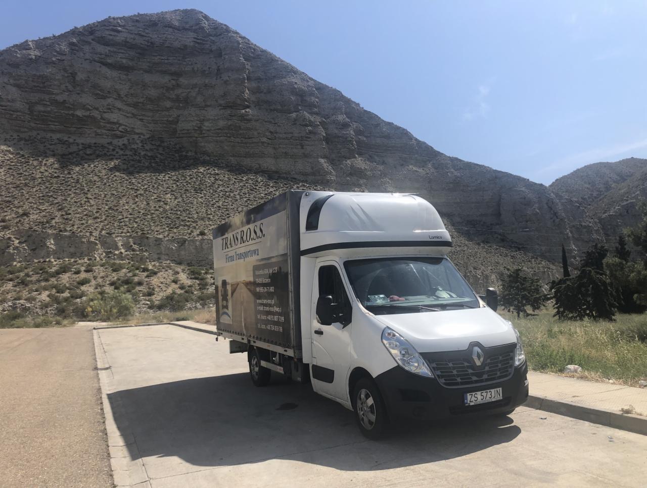 Biały bus firmy transportowej Trans R.O.S.S. zaparkowany na betonowym placu z widokiem na skaliste wzgórza w słoneczny dzień.