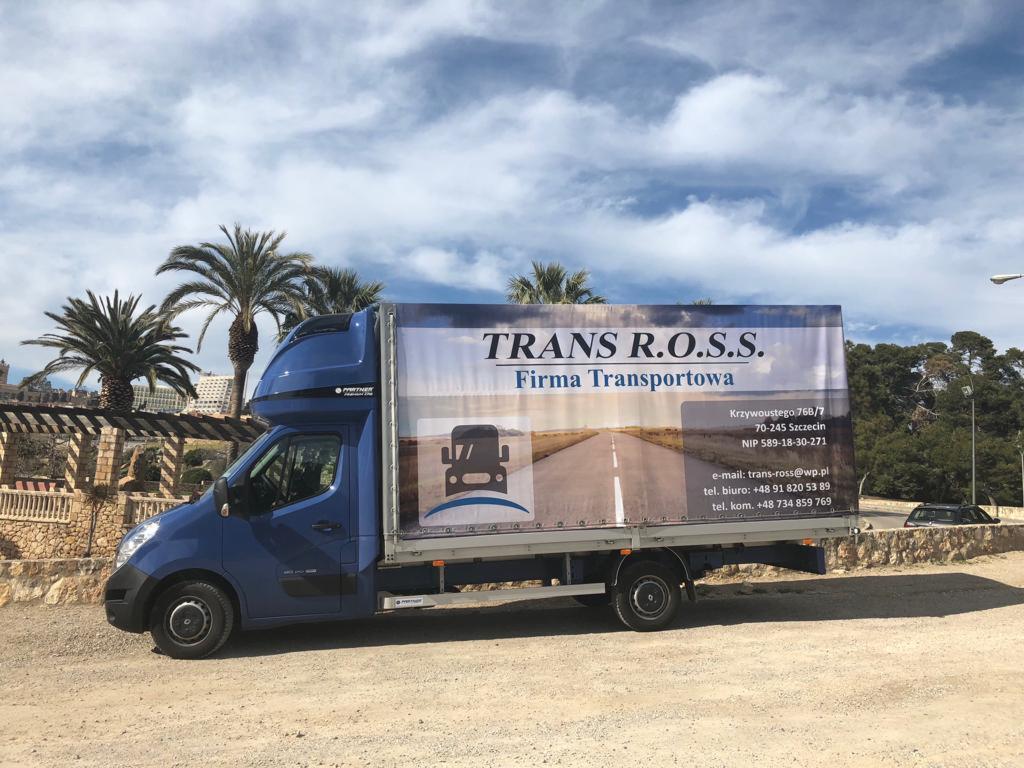 Niebieski bus transportowy firmy TRANS R.O.S.S. z reklamą na plandece, zaparkowany na żwirowej nawierzchni z palmami w tle.