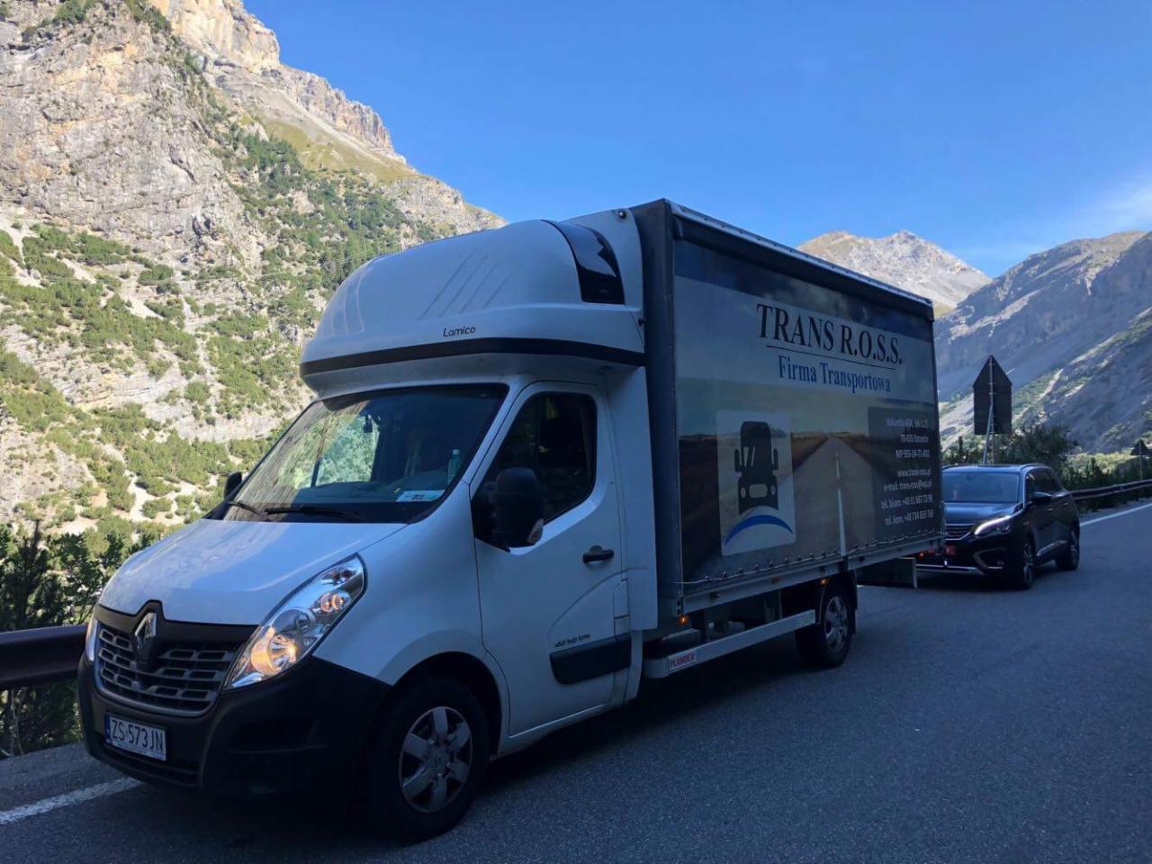 Biały bus dostawczy marki Renault z zabudową Lamico, z logo firmy Trans R.O.S.S., zaparkowany na poboczu drogi górskiej, w tle widok na Alpy.