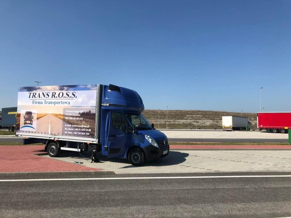 Niebieski bus dostawczy z plandeką, na której widnieje logo firmy transportowej Trans R.O.S.S., zaparkowany na parkingu z widocznymi w tle innymi pojazdami ciężarowymi i błękitnym niebem.