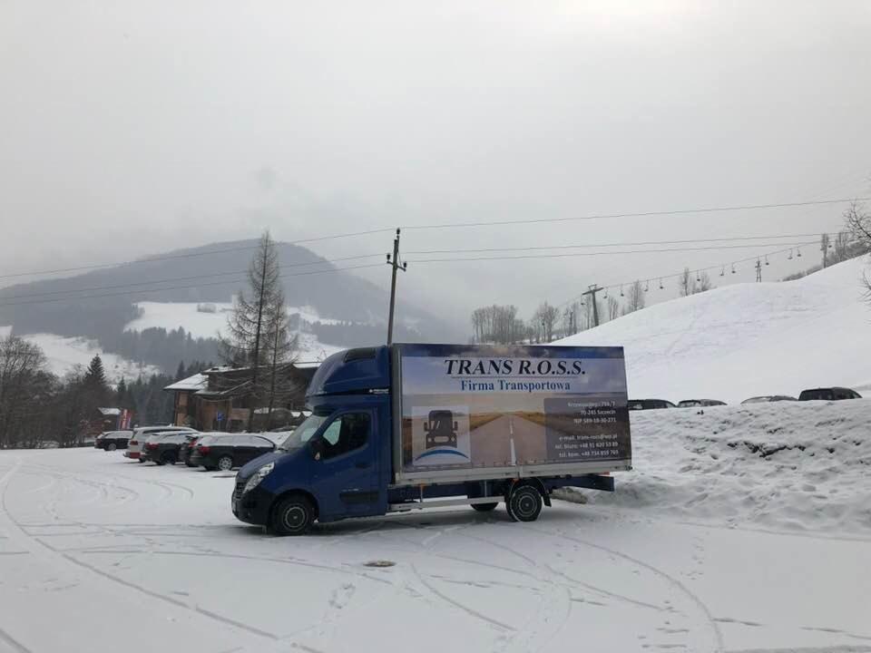 Niebieski bus dostawczy firmy transportowej TRANS R.O.S.S. zaparkowany na zasypanym śniegiem parkingu, w tle góry i wyciąg narciarski w pochmurny dzień.