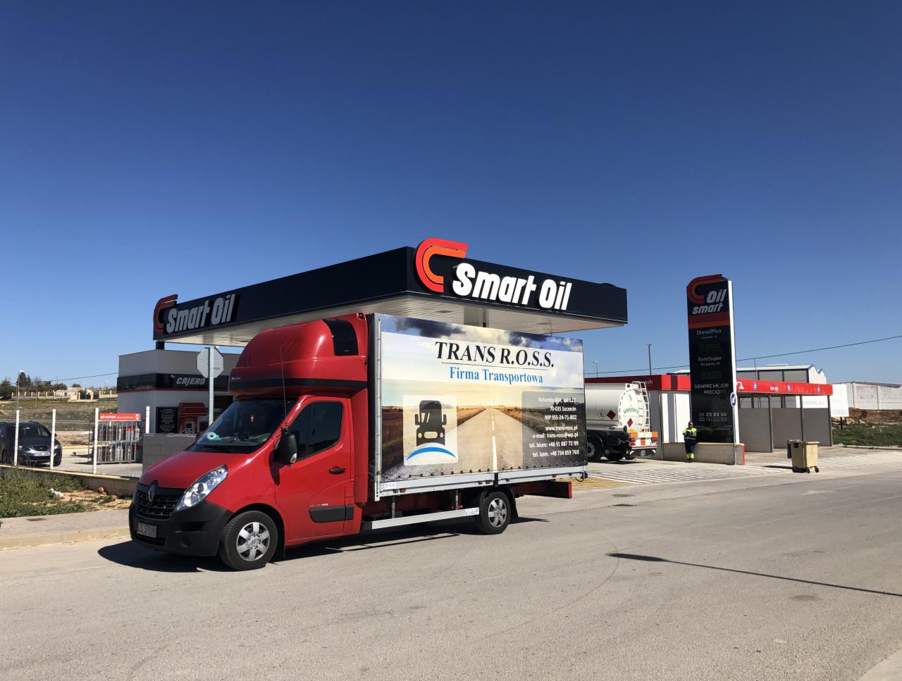 Czerwony bus transportowy firmy TRANS R.O.S.S. z reklamą na plandece, zaparkowany na stacji benzynowej Smart Oil w słoneczny dzień.