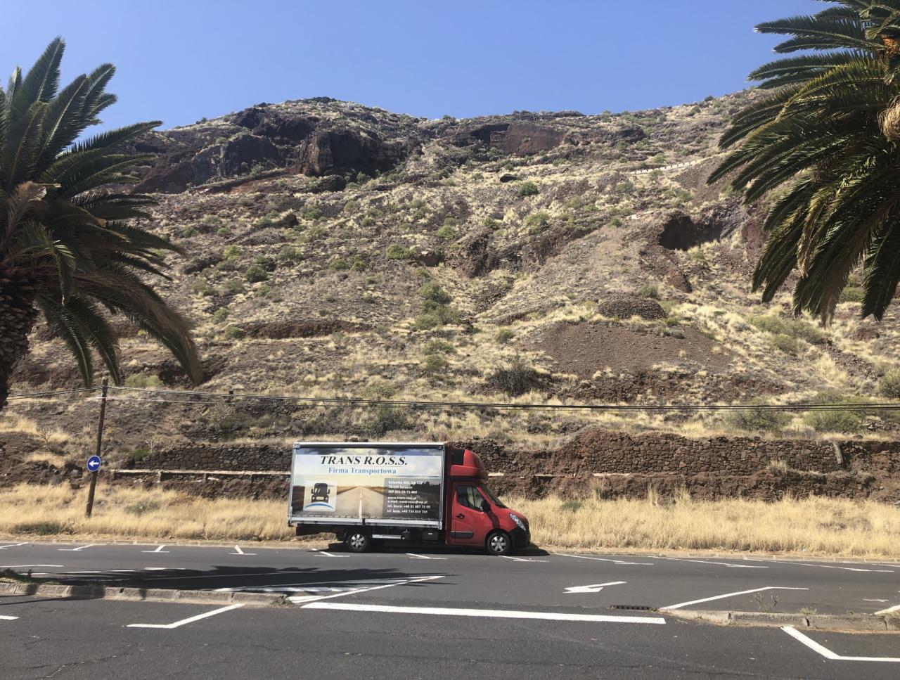 Czerwony bus transportowy 'TRANS R.O.S.S. Firma Transportowa' zaparkowany na parkingu z widokiem na zbocze góry i palmy.