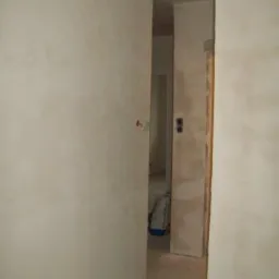 Ściany w trakcie wygładzania gładzią szpachlową, widoczny fragment instalacji elektrycznej i worek z materiałami budowlanymi w tle.