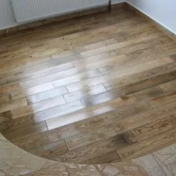 Ukończona podłoga z drewnopodobnych paneli ułożonych w jodełkę, z marmurowym wykończeniem przy ścianach i zaokrąglonym przejściem do innej części podłogi.