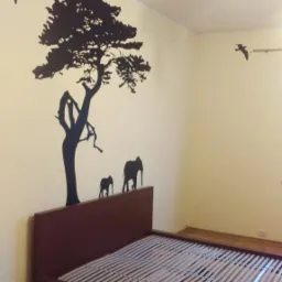 Ściana pokoju z naklejką przedstawiającą afrykański krajobraz: drzewo, słonie i ptaki, nad brązowym łóżkiem ze stelażem.