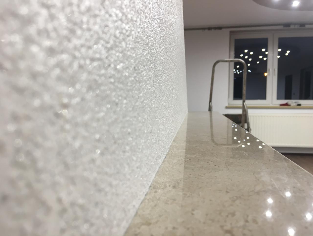 Błyszcząca, biała ściana z efektem dekoracyjnym, widoczny blat i drabina w tle, oświetlenie sufitowe odbijające się w blacie.