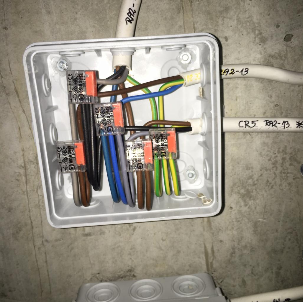 Otwarta, biała puszka elektryczna z widocznymi przewodami w różnych kolorach połączonych pomarańczowymi złączkami Wago, zamontowana na szarej ścianie.