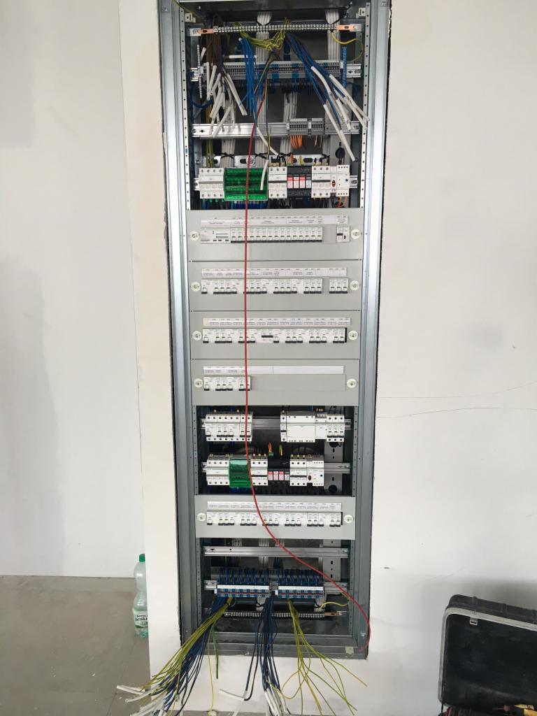 Rozdzielnica elektryczna w trakcie montażu w surowym wnętrzu, widoczne liczne kable i zabezpieczenia, skrzynka narzędziowa w tle.