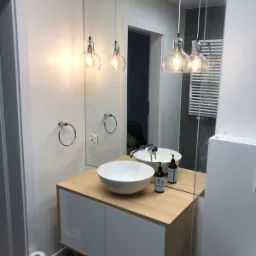 Nowoczesna łazienka z okrągłą białą umywalką nablatową na szafce z drewnianym blatem i białymi frontami, oświetlona trzema szklanymi lampami wiszącymi w kształcie żarówek, z dużym lustrem...