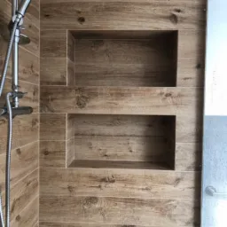 Wykończona ściana prysznica z dwoma wnękami, pokryta płytkami imitującymi drewno. Widoczna chromowana bateria prysznicowa i fragment szklanej kabiny.