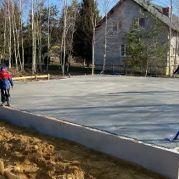 Wylany fundament płyty betonowej pod dom jednorodzinny, dwóch chłopców spaceruje po powierzchni, w tle drzewa i budynek mieszkalny.