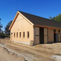 Nowo wybudowany budynek gospodarczy z jasnego drewna, z ciemnym dachem, otwartymi drewnianymi wrotami i małymi oknami, na placu budowy z widocznymi śladami opon.