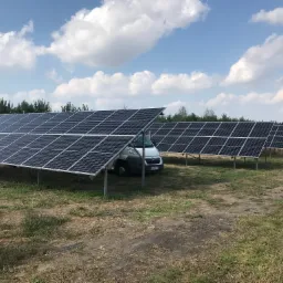 Rząd paneli słonecznych na metalowych podporach, częściowo zasłaniający białego busa na tle błękitnego nieba z białymi chmurami i zielonych drzew.