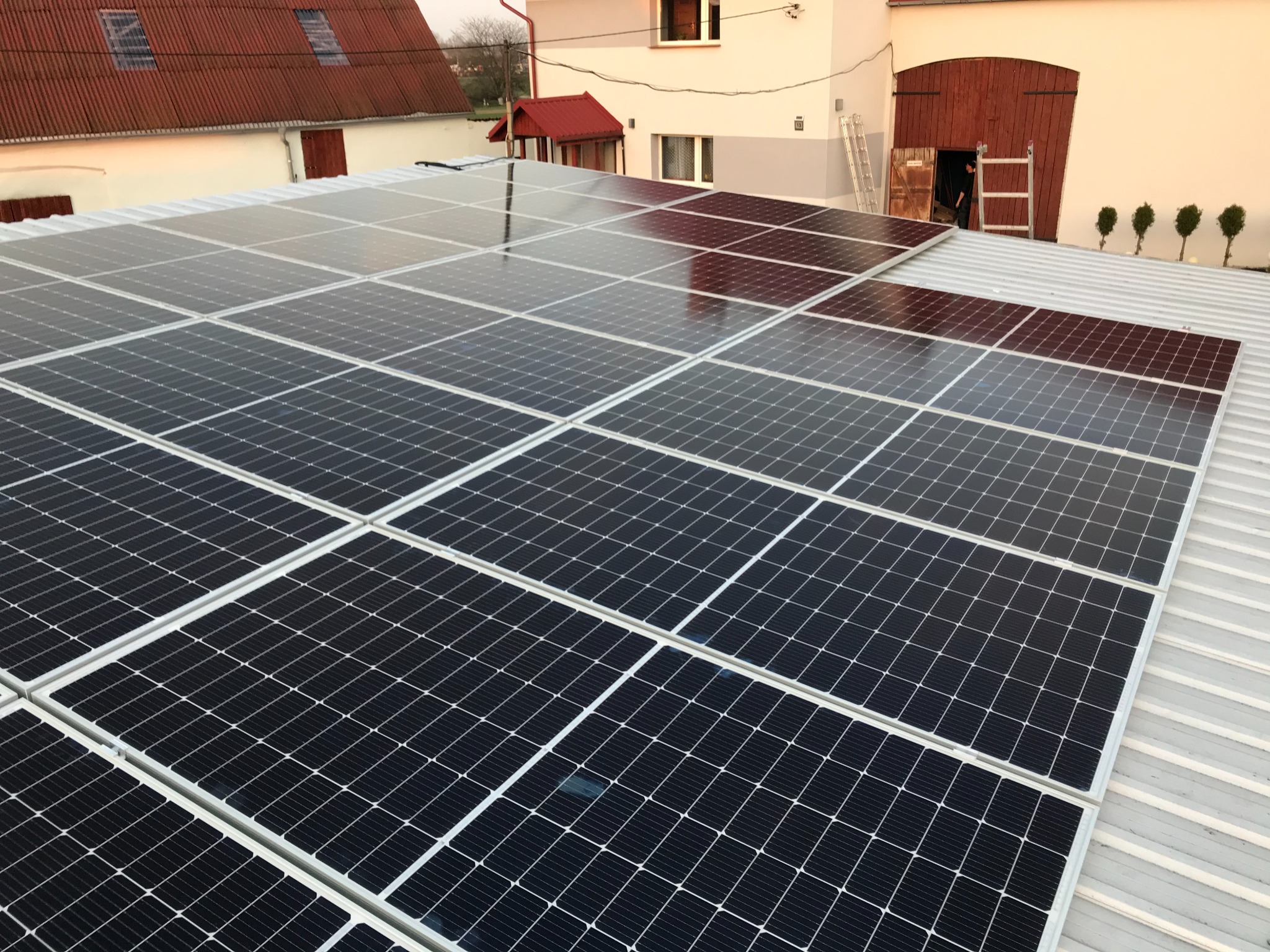 Instalacja paneli słonecznych na blaszanym dachu budynku gospodarczego, widoczna drabina i otwarte drzwi do budynku.