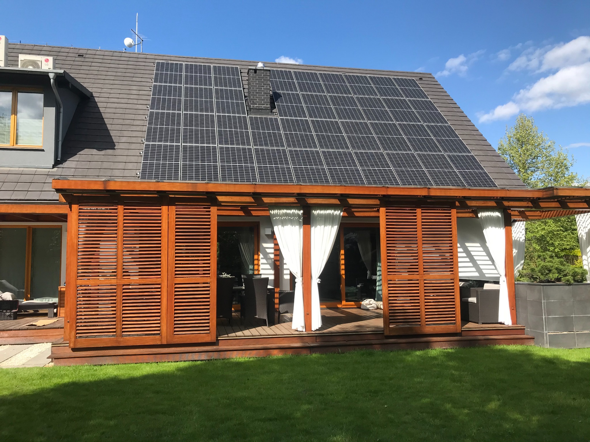 Dom z szarym dachem i panelami słonecznymi, widok z ogrodu na drewniany taras z przesuwnymi żaluzjami i białymi zasłonami. Na dachu widoczny komin i antena.