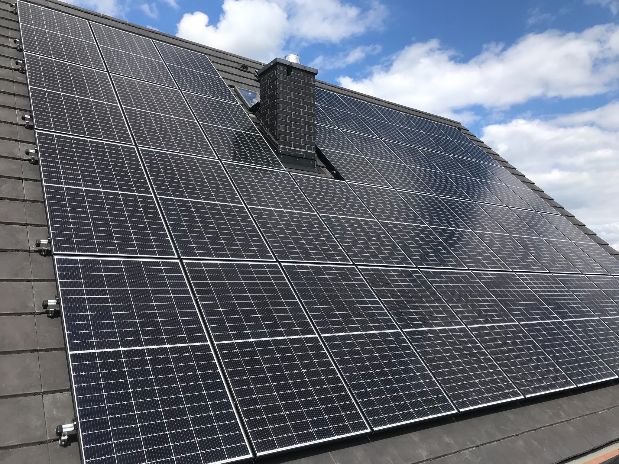 Panele słoneczne na dachu z ciemnej dachówki, widoczny komin z ciemnej cegły i fragment błękitnego nieba z chmurami.