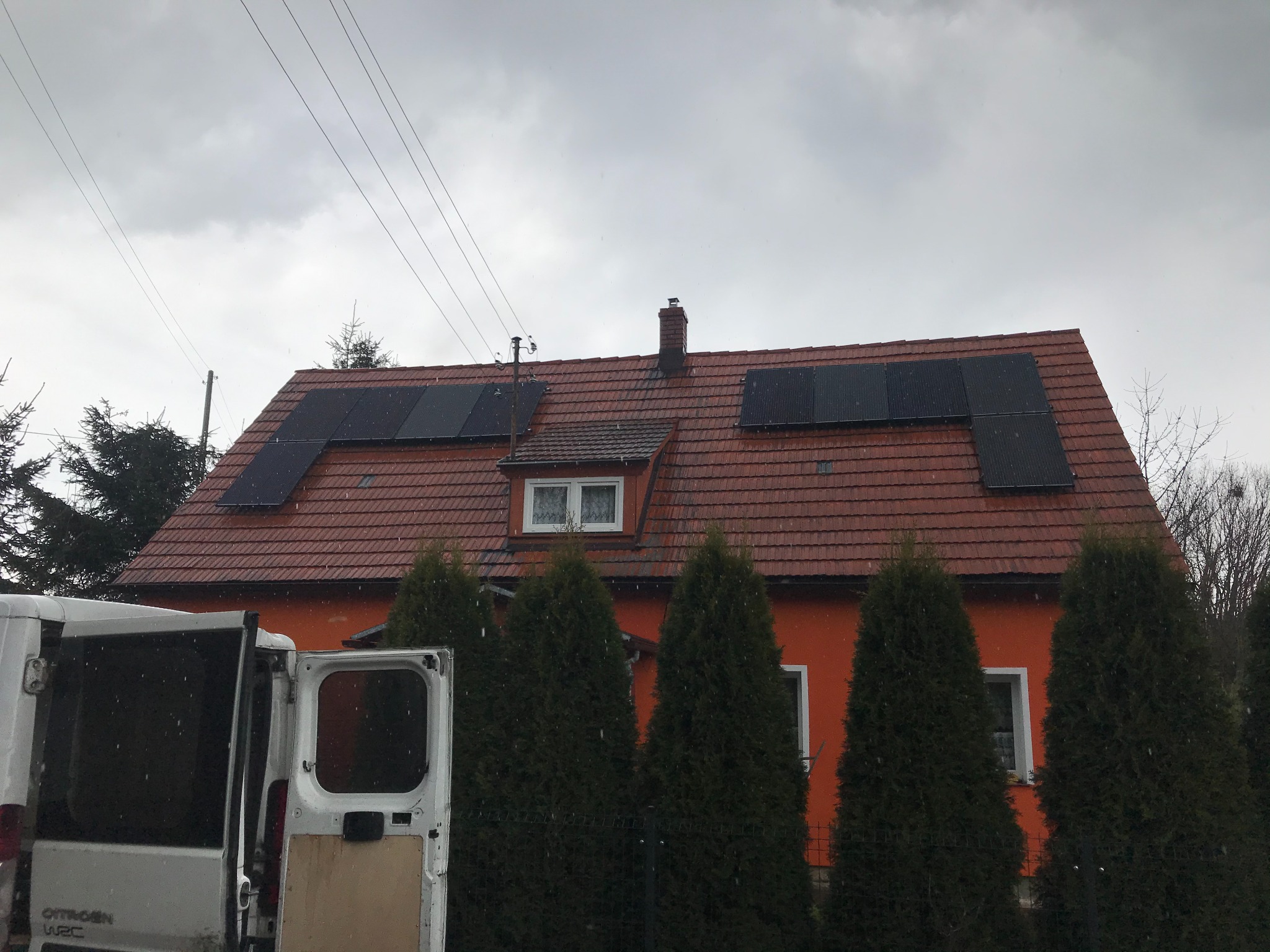 Pomarańczowy dom jednorodzinny z czerwoną dachówką, na której zainstalowane są czarne panele słoneczne. Z boku widoczny biały samochód dostawczy z otwartymi drzwiami, a w tle pochmurne niebo.