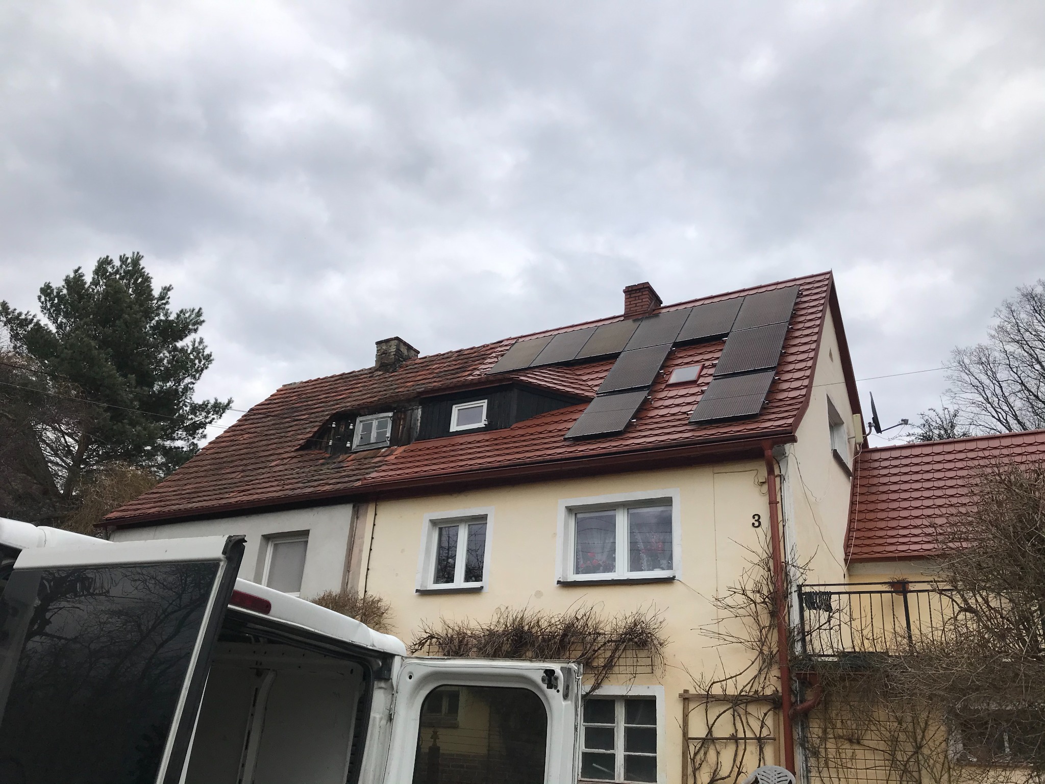 Dom jednorodzinny z panelami słonecznymi na czerwonym dachu, widoczne otwarte drzwi białego samochodu dostawczego, pochmurne niebo.