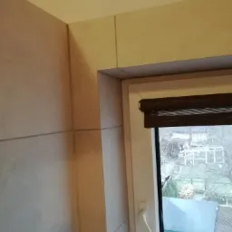 Wykończona ściana w beżowych płytkach ceramicznych przy oknie z brązowymi roletami, widok na zewnątrz na szary ogród.