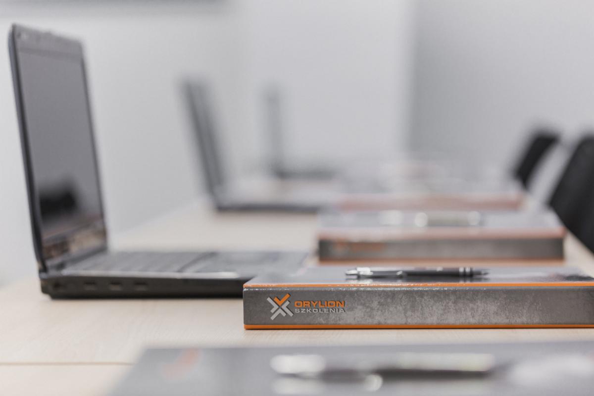 Sala szkoleniowa: laptopy i materiały z logo firmy szkoleniowej Orylion Szkolenia, długopis na materiałach.