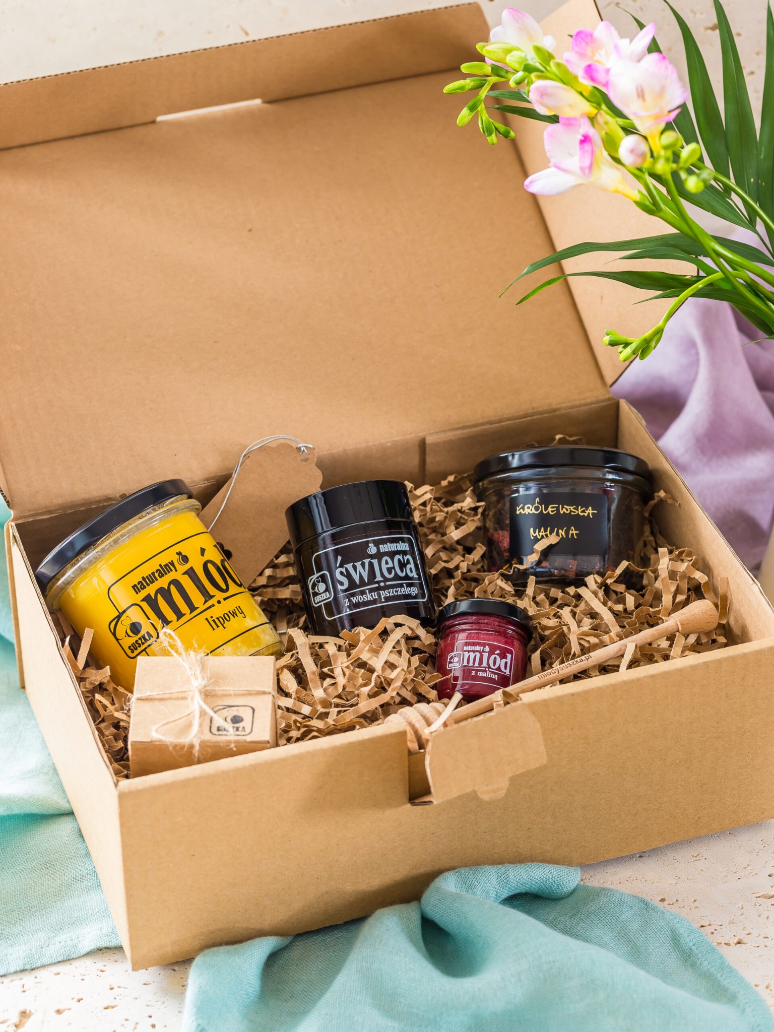 Otwarty kartonowy kosz prezentowy wypełniony papierowym wypełniaczem, zawierający słoiki z miodem o różnych smakach, świecę z wosku pszczelego i mały prezent zapakowany w papier, obok bukiet frezji...