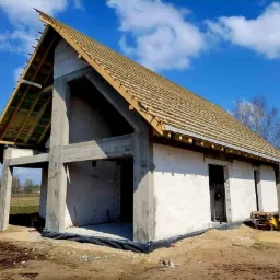 Surowy stan domu z betonu komórkowego z częściowo wykonanym drewnianym dachem i widocznymi elementami konstrukcyjnymi.