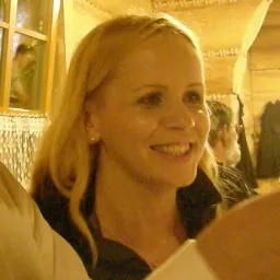 Portret uśmiechniętej blondynki z perłą w uchu, w tle okno z odbiciem i fragment wnętrza restauracji.