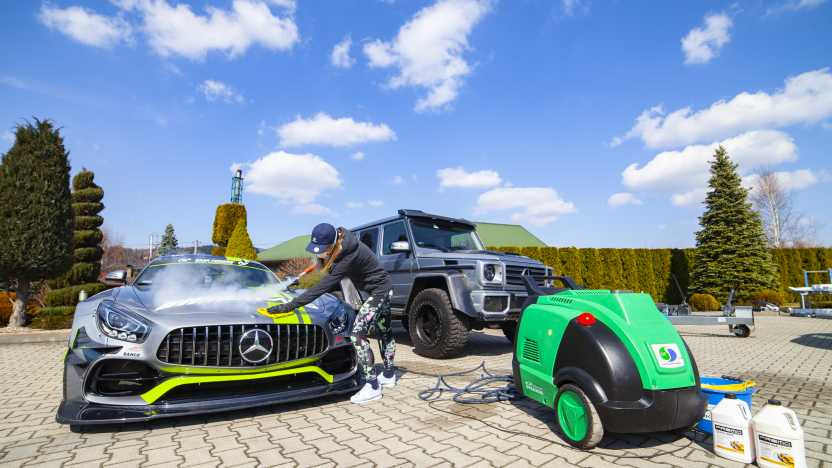 Mycie parowe srebrnego sportowego Mercedesa AMG z zielonymi akcentami przez osobę w czarnej bluzie i wzorzystych legginsach, w tle szary Mercedes G-klasy i zielona myjka parowa na kostce brukowej.