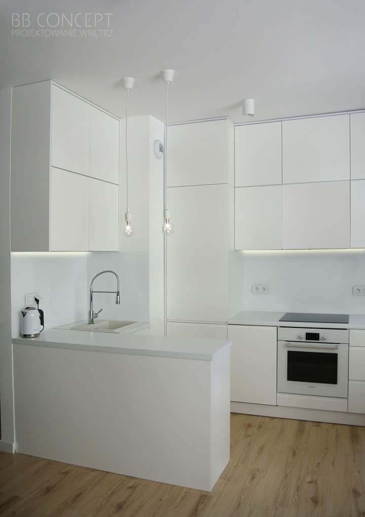 Minimalistyczna, biała kuchnia z drewnianą podłogą, oświetleniem LED pod szafkami wiszącymi i wiszącymi lampami z żarówkami Edisona.