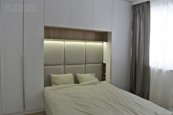 Nowoczesna sypialnia z zabudową meblową: białe szafy wnękowe zintegrowane z tapicerowanym zagłówkiem podświetlonym listwą LED, szare zasłony przy oknie.