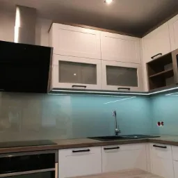 Nowoczesna kuchnia z białymi szafkami, drewnianymi blatami i turkusowym szkłem na ścianie, oświetlona listwą LED pod szafkami wiszącymi.