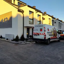 Biały bus firmy budowlanej z reklamą tynków maszynowych i ociepleń zaparkowany przed nowoczesnymi szeregowcami w promieniach zachodzącego słońca.