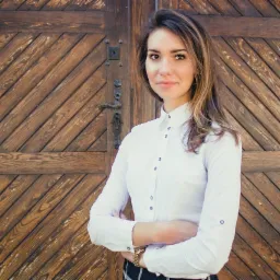 Portret kobiety w białej koszuli stojącej na tle rustykalnych drewnianych drzwi z ozdobnym uchwytem.