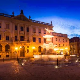 Nocny widok na oświetloną fontannę z rzeźbą na tle klasycystycznej architektury miejskiej i granatowego nieba.