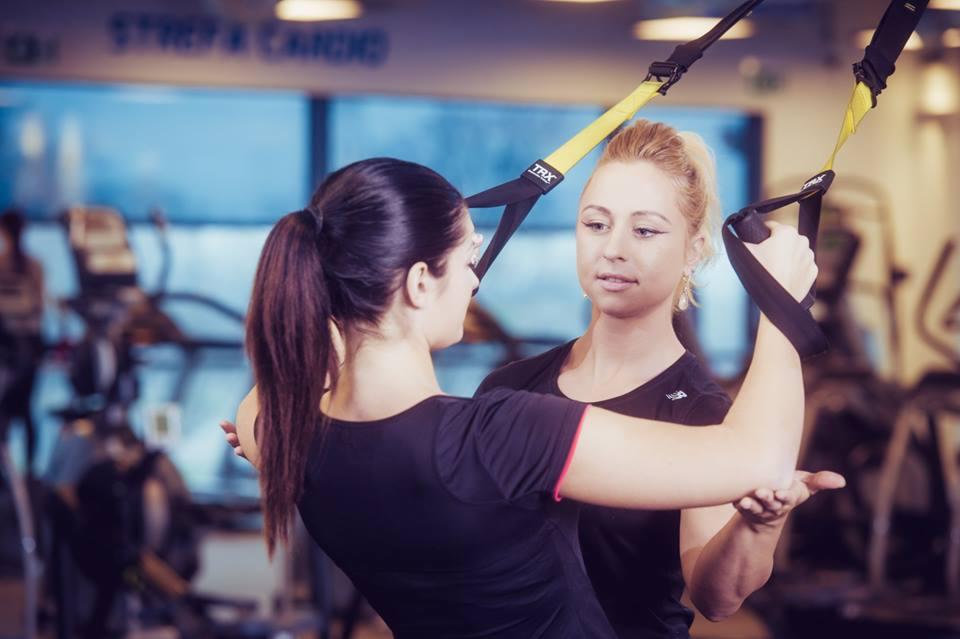Kobieta ćwiczy z użyciem systemu TRX pod okiem instruktorki w siłowni, widoczne urządzenia do ćwiczeń w tle.